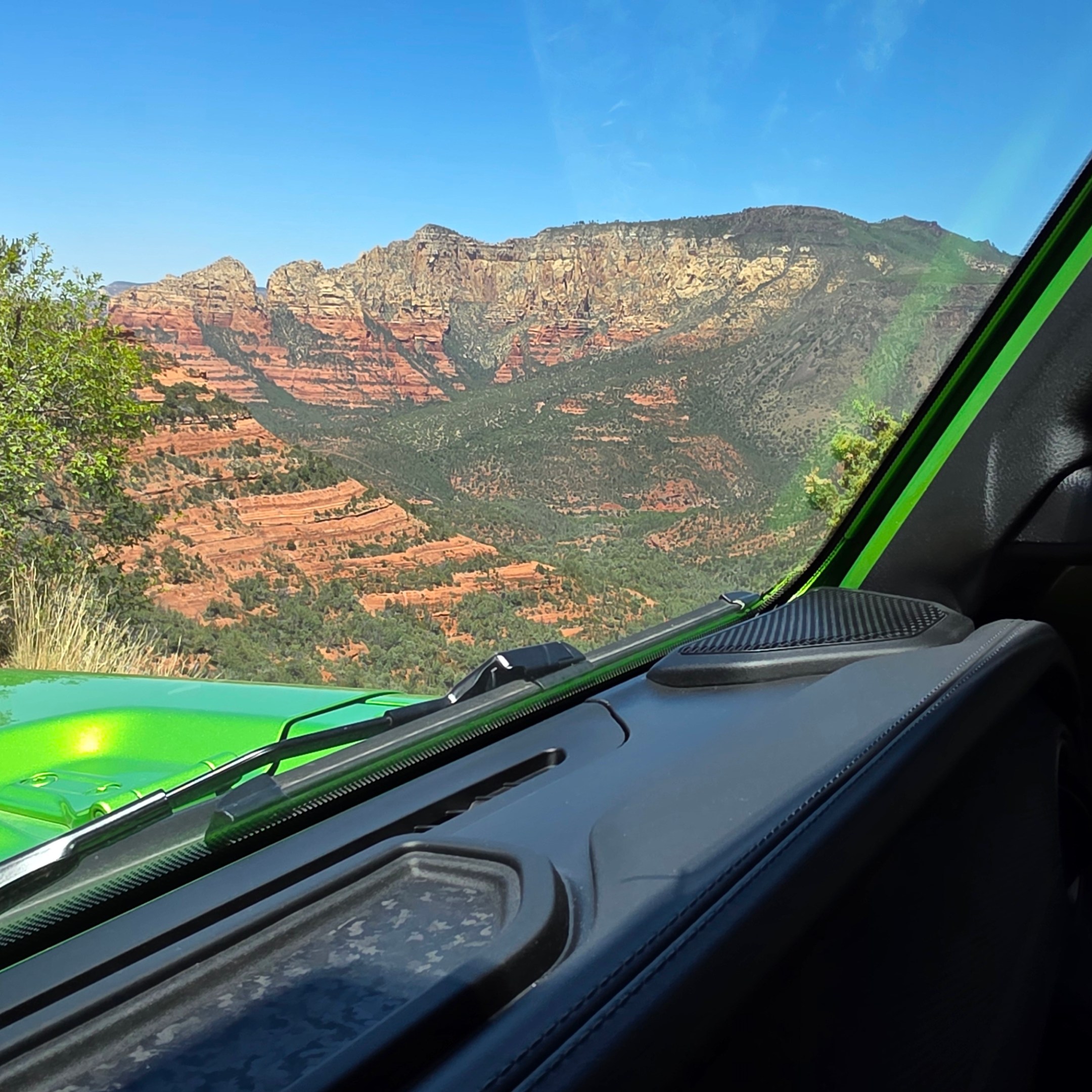 View from a green car's interior of red rock mountains and trees under a clear blue sky.
