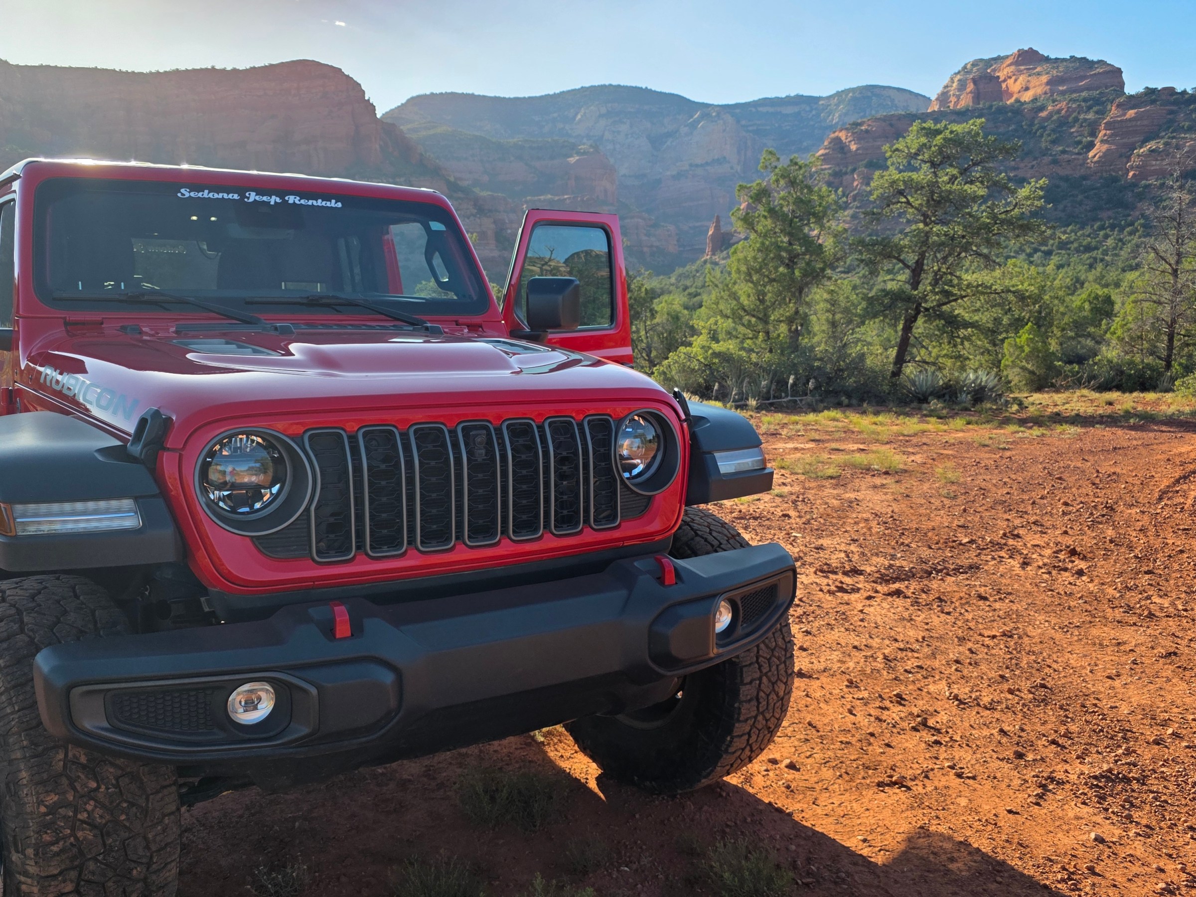 Red Jeep on dirt road near rocky landscape and trees.