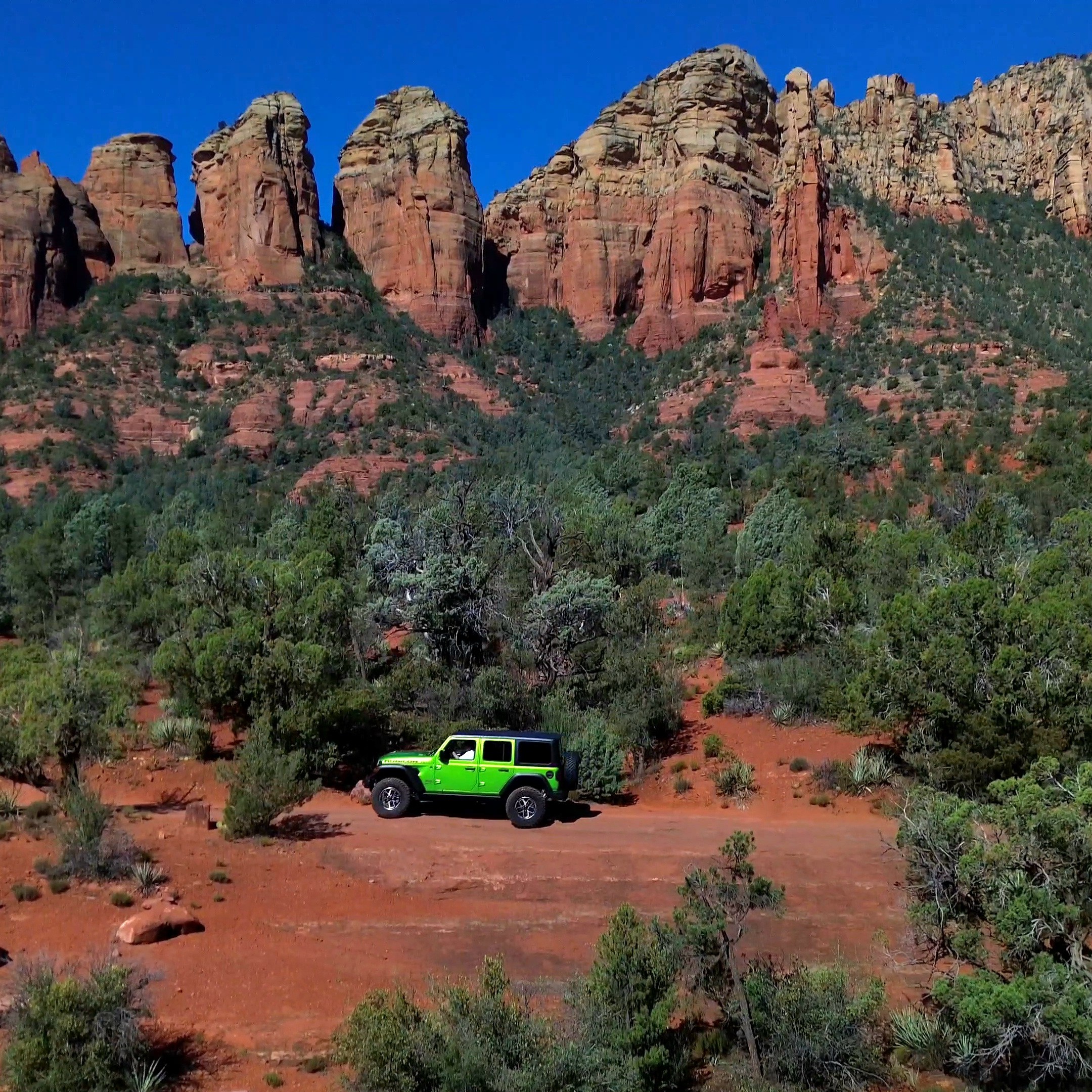 Green jeep on red desert terrain with rocky cliffs in the background.
