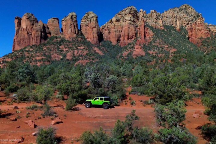 Green jeep on red desert terrain with rocky cliffs in the background.