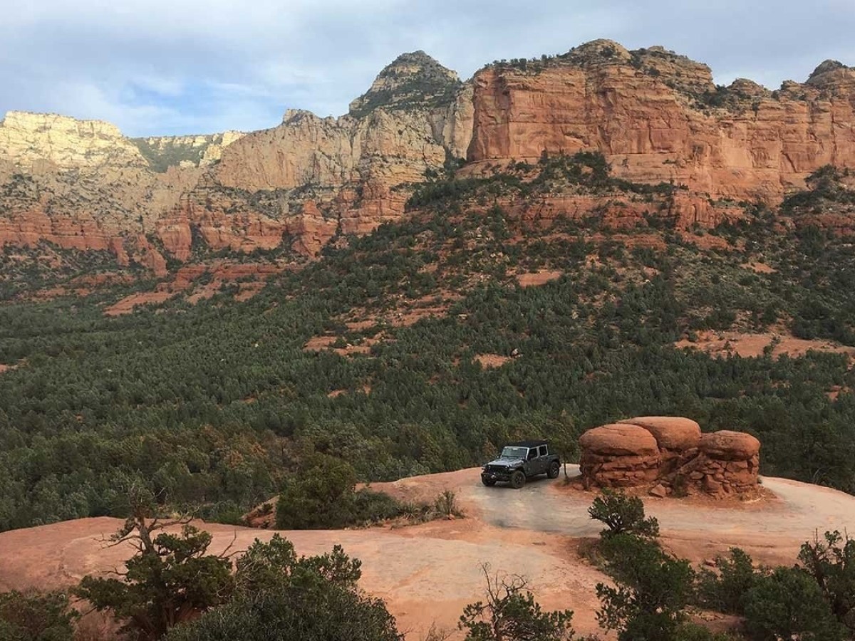 Jeep on rocky terrain with red cliff backdrop and green forest.