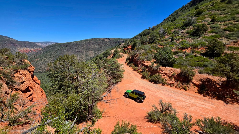 Green jeep on a dirt mountain road surrounded by trees and hills.