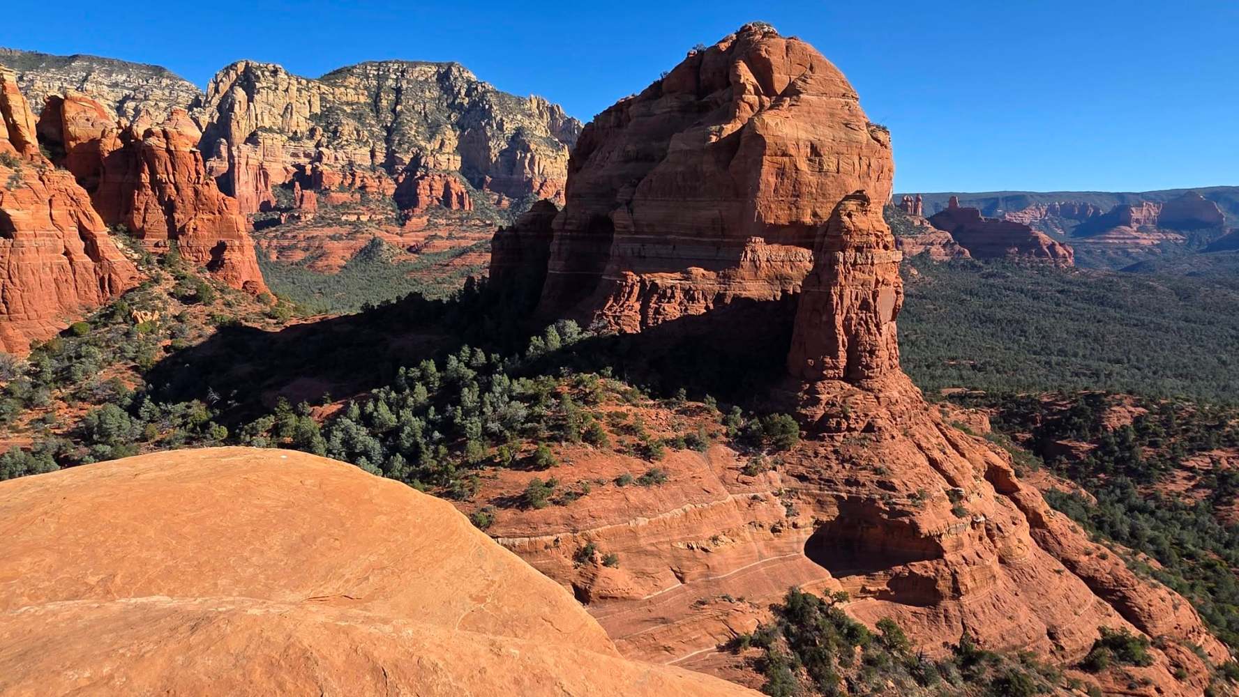 Red rock formations under a clear blue sky at Sedona, Arizona with distant cliffs and trees.