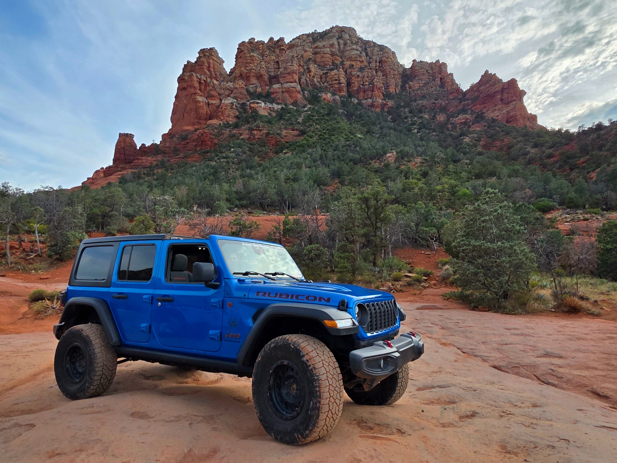 Blue jeep on desert terrain with rocky cliffs in the background.