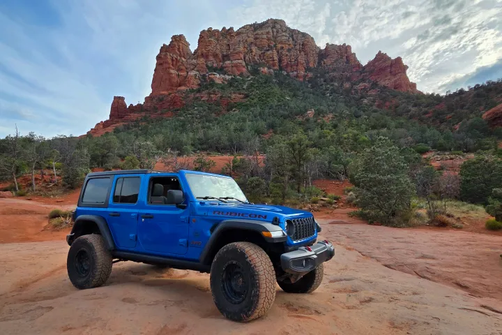 Blue jeep on desert terrain with rocky cliffs in the background.