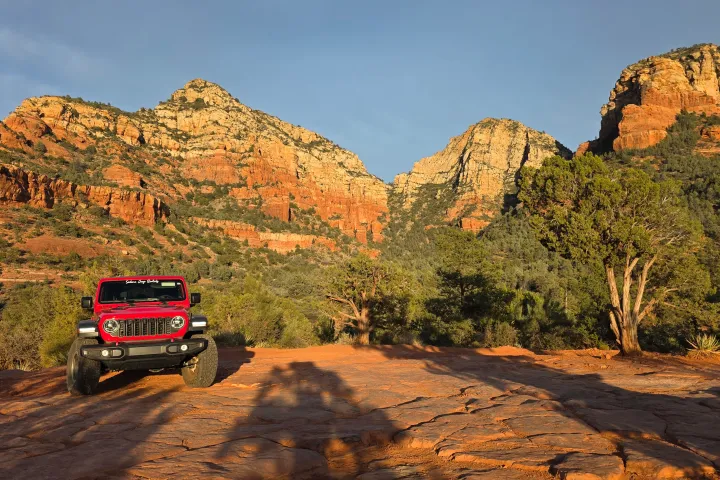 Red SUV on rocky terrain with red cliffs and greenery in the background.