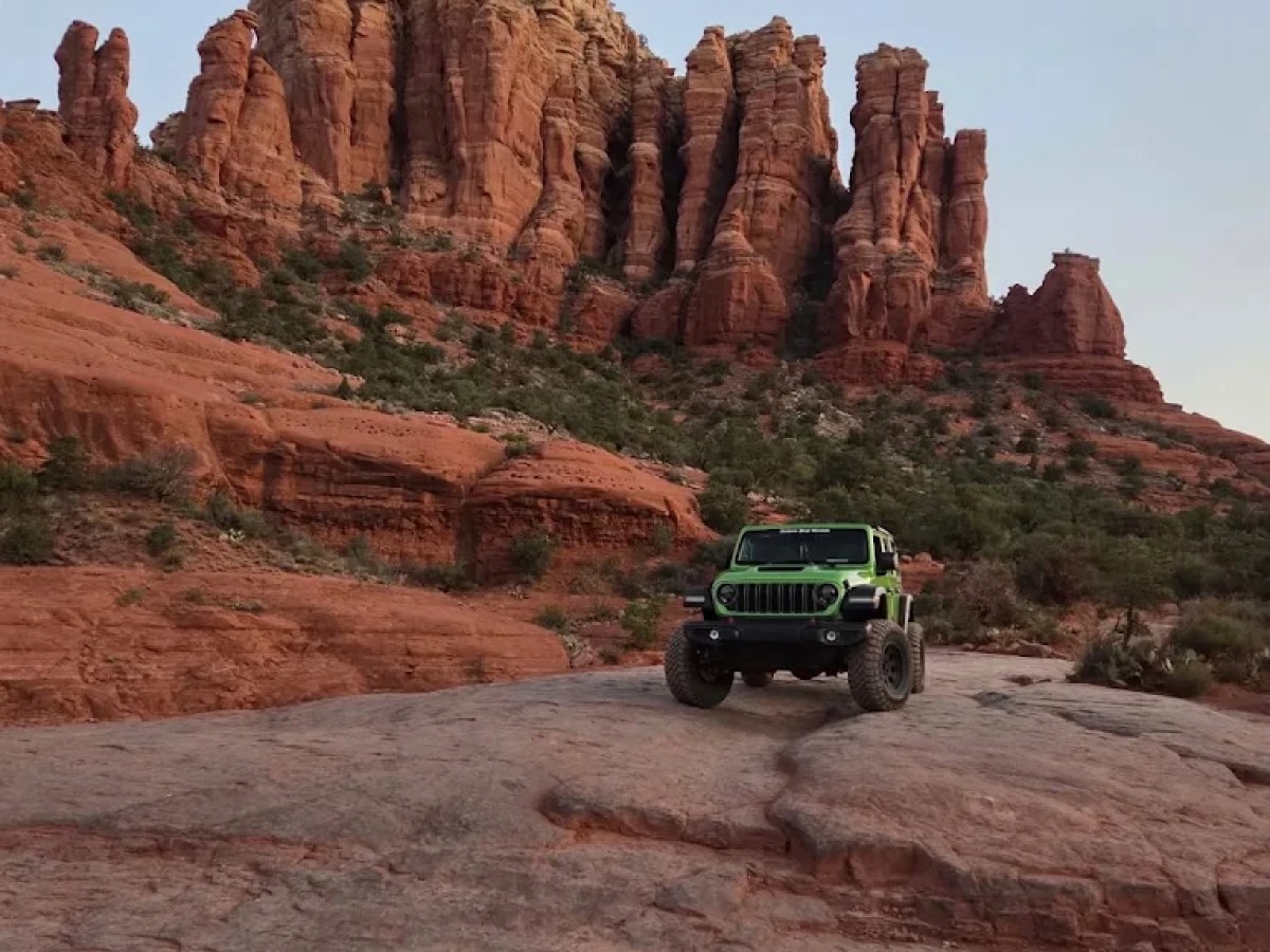 Green off-road vehicle on rocky terrain with large red rock formations in the background.