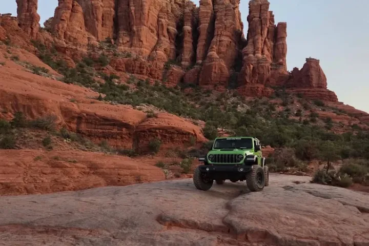 Green off-road vehicle on rocky terrain with large red rock formations in the background.
