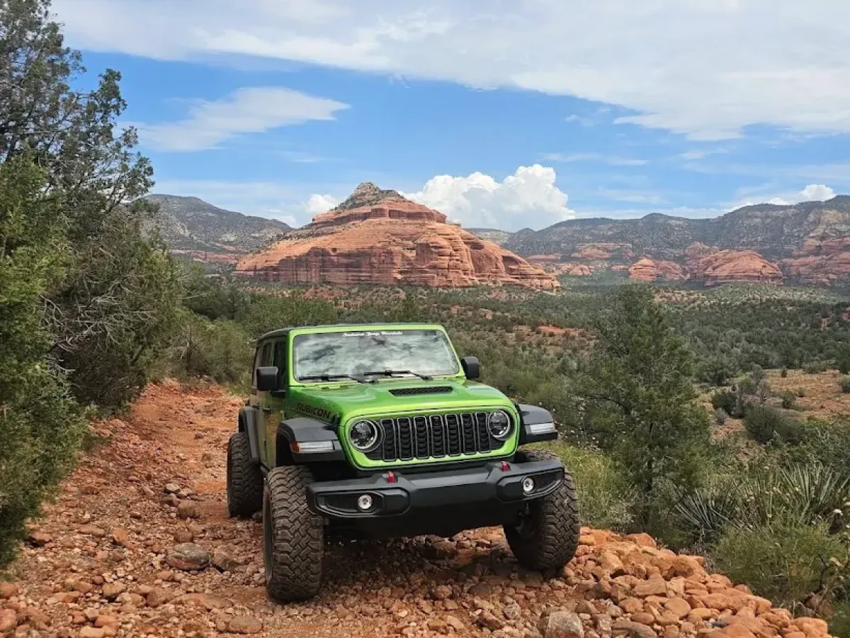 Green off-road vehicle on rocky trail with red rock formations in the background.
