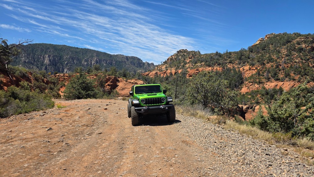 Green off-road vehicle on a dirt trail in a rocky, hilly landscape under a blue sky.