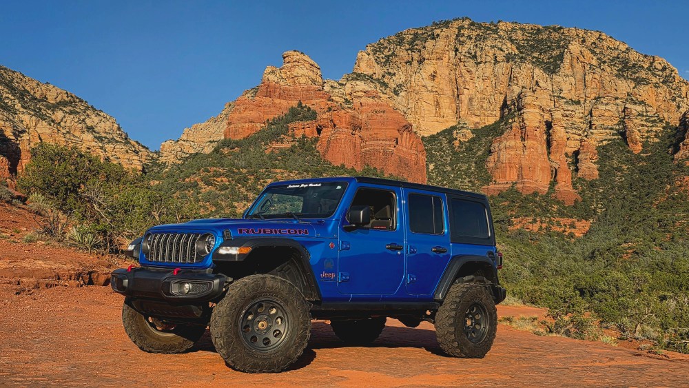 A blue Jeep on a desert landscape with rocky red cliffs in the background.