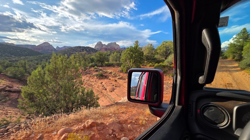 View from a car window of a rocky, desert landscape with trees and mountains under a cloudy sky.