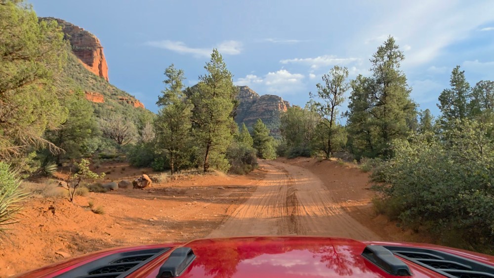 Red vehicle driving on a dirt road in a desert landscape with trees and mountains.