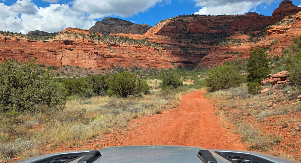 Red dirt road through canyon with rocky cliffs and sparse greenery under blue sky.