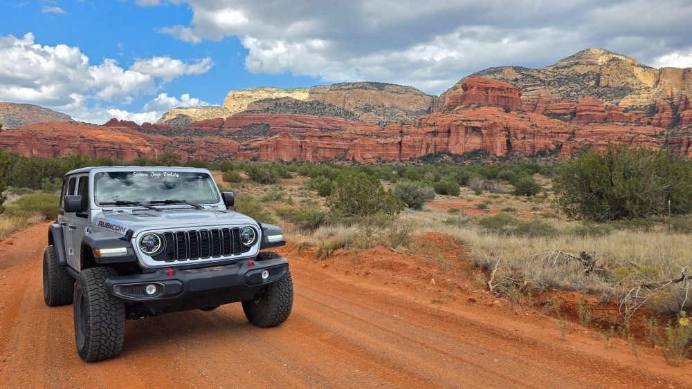 SUV on dirt road with red rock formations and cloudy sky in background.